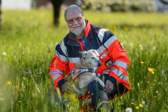 Rettungshund sucht im Wald nach einer versteckten Person, die Hundeführerin beobachtet die Arbeit
